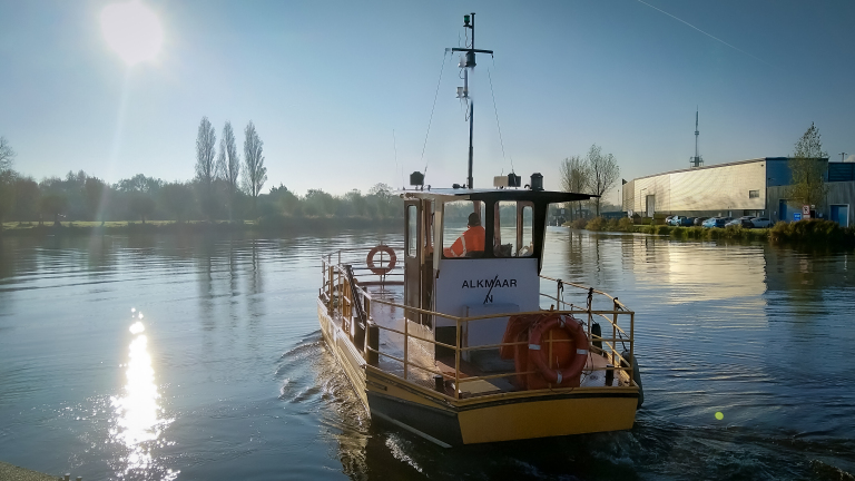 Kleine veerboot met de naam "Alkmaar" op een rustige rivier, bij zonsopgang.