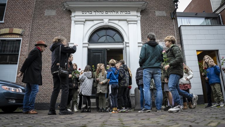 Een groep mensen, waaronder kinderen met bloemen, verzamelt zich voor de ingang van een gebouw met Hebreeuwse tekst boven de deur.