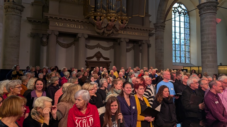 Groep mensen verzameld in een kerk, met een pijporgel en gebrandschilderd raam op de achtergrond.