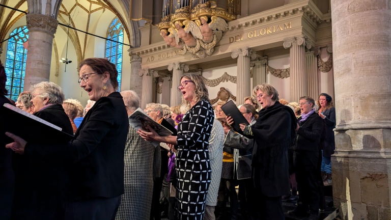 Koor zingt in een kerk met decoratieve zuilen en glas-in-loodramen. Op achtergrond staat een orgel met tekst "AD SOLI DEI GLORIAM".