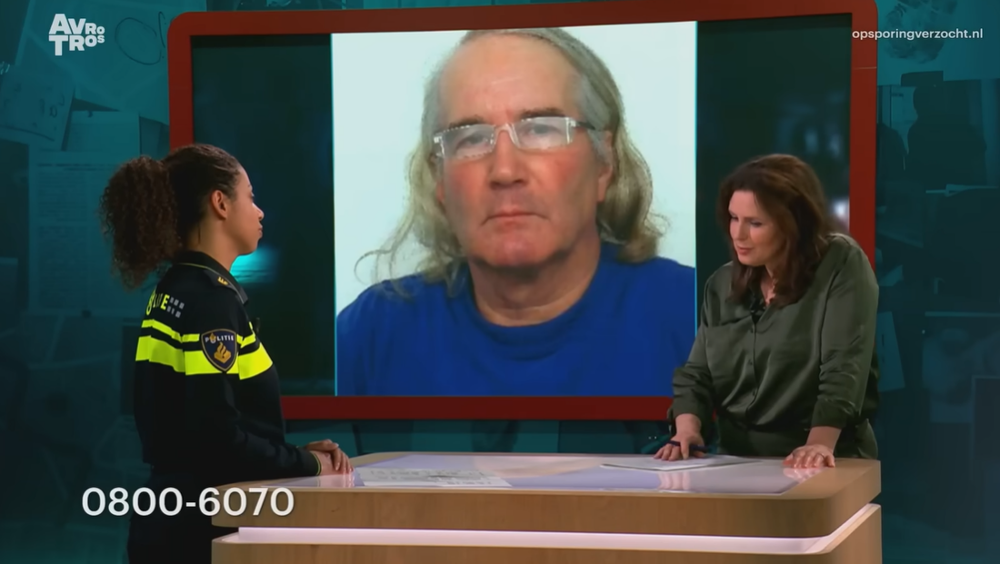 Twee vrouwen staan in een televisiestudio, een daarvan draagt een politie-uniform. Op de achtergrond is een foto van een oudere man met lang haar en een bril te zien.