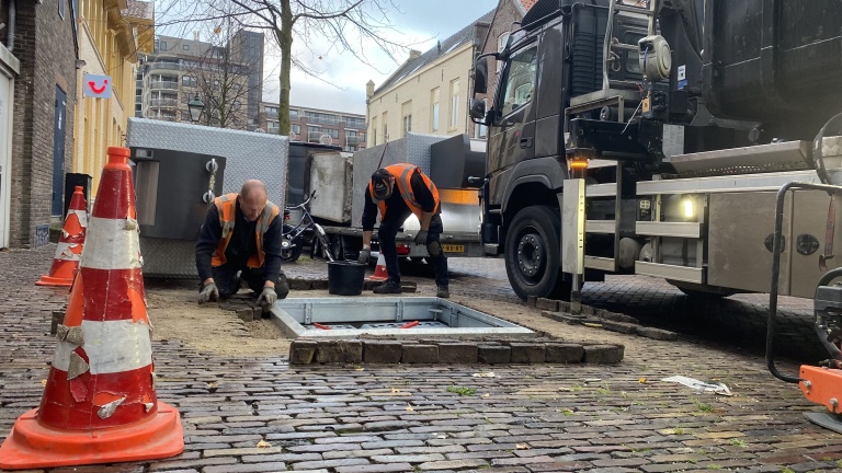 Twee wegwerkers in oranje veiligheidsvesten werken aan een installatie in de straat, omgeven door verkeerskegels en een vrachtwagen.