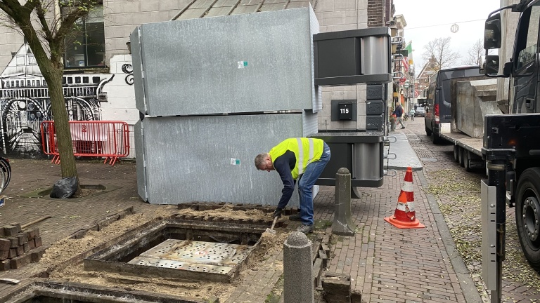 Man in geel veiligheidsvest werkt op straat naast grote metalen structuren en verkeerskegel.