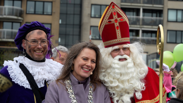 Sinterklaas en Zwarte Piet poseren met een vrouw, allemaal glimlachend, voor een gebouw op de achtergrond.