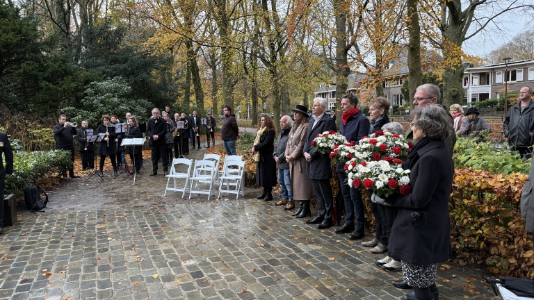 Mensen staan in een park bij een herdenkingsceremonie, sommigen houden bloemen vast; een blaasorkest speelt op de achtergrond.