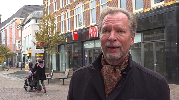 Midden op een winkelstraat staat een man met een jas en sjaal, terwijl op de achtergrond mensen voorbij lopen.