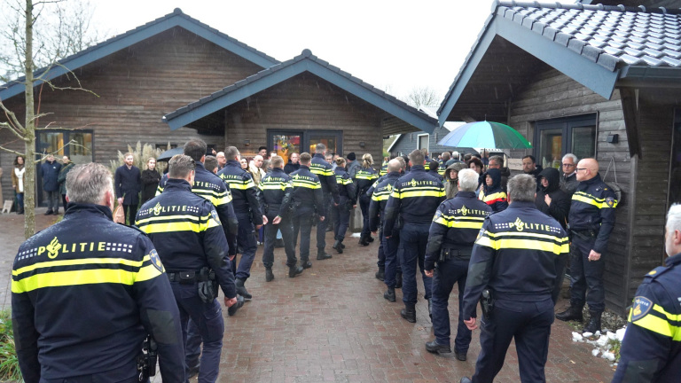 Politieagenten staan buiten in de regen bij houten gebouwen, omgeven door mensen.