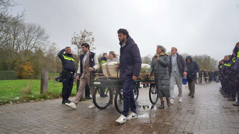 Mensen lopen met een kist op een handkar, terwijl politieagenten een erehaag vormen.