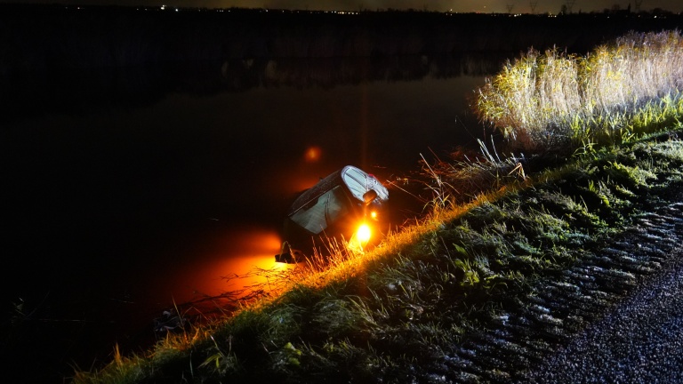 Auto half onder water in een sloot bij nacht, met de koplampen nog aan en verlichte graskant.