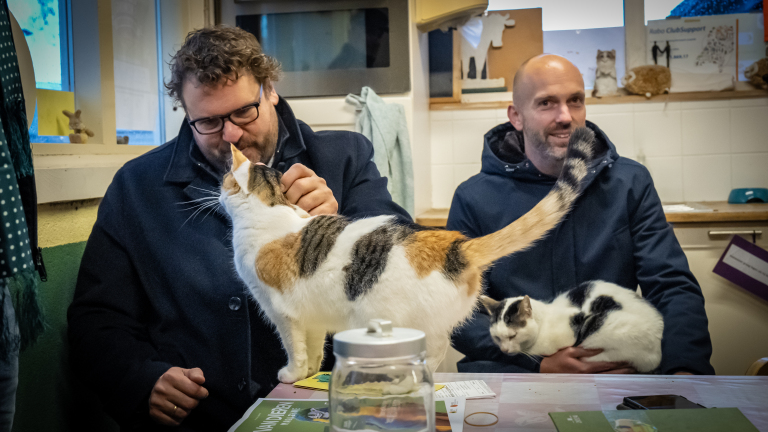 Twee mannen zitten binnen aan tafel met katten op schoot.