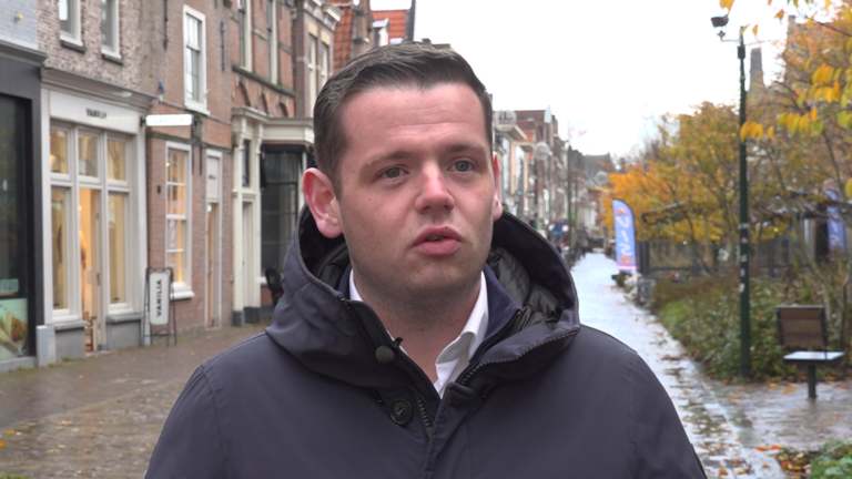 Man in een jas staat in een herfstachtige straat met winkels en bomen.