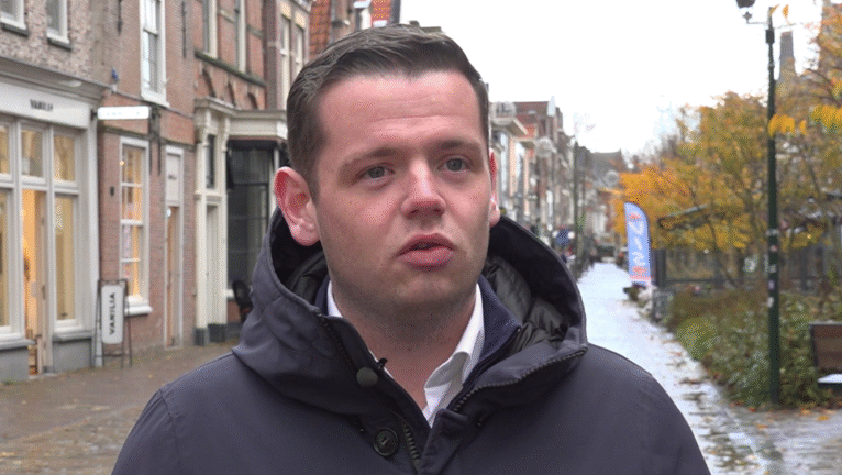 Man in een jas staat in een herfstachtige straat met winkels en bomen.