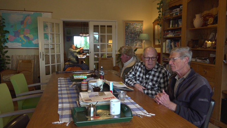 Twee mannen zitten aan een tafel in een kamer met boekenplanken en een kaart van Europa aan de muur; op de achtergrond praten mensen in een andere kamer.