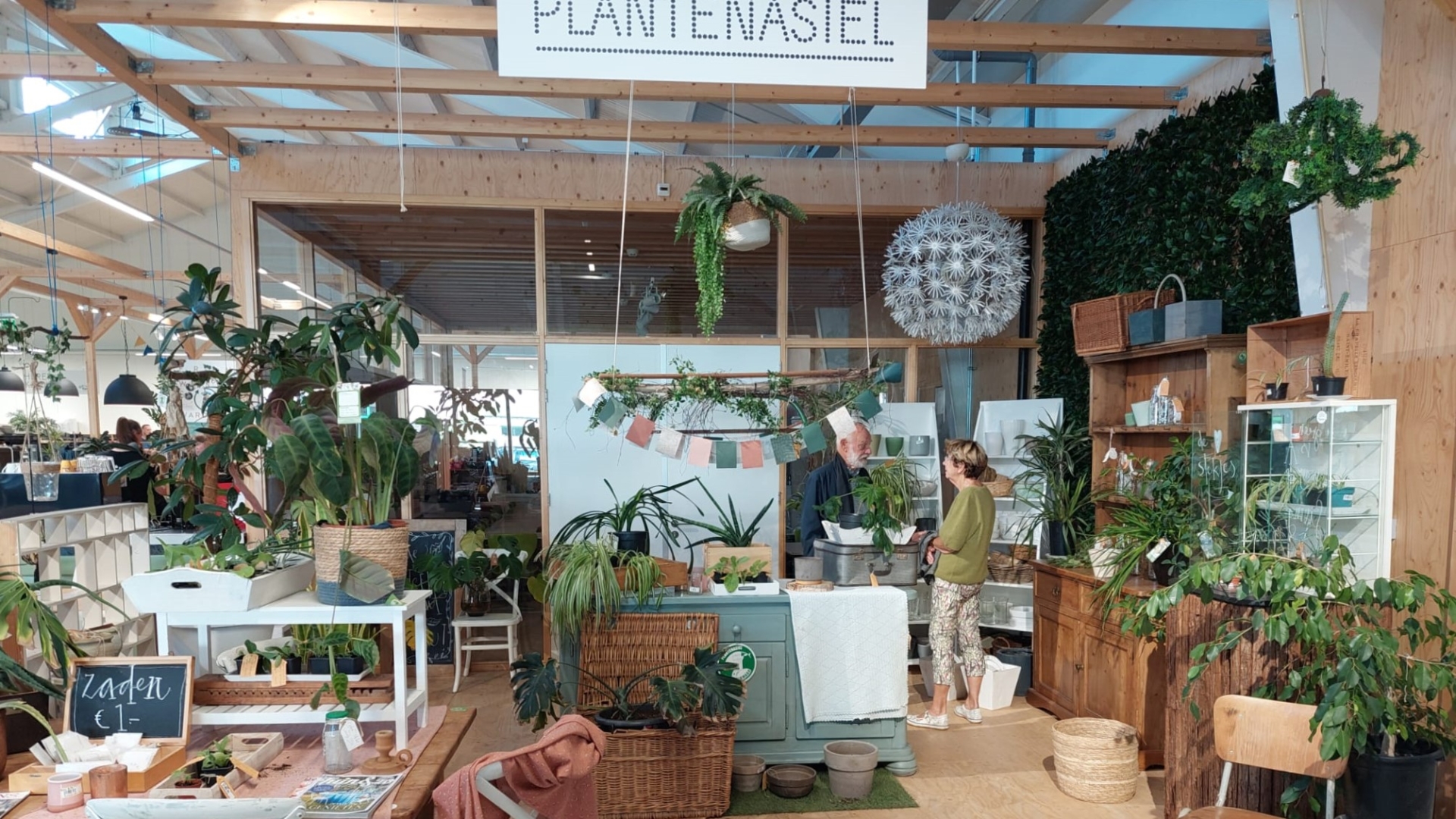 Binnenruimte van een plantenwinkel met diverse planten en houten meubels, grote hanglamp en mensen die met elkaar praten.