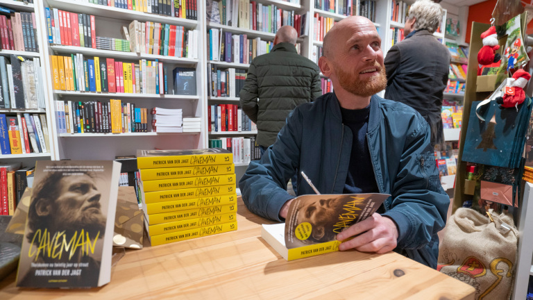 Een man signeert een boek in een boekwinkel, met een stapel van hetzelfde boek op tafel.