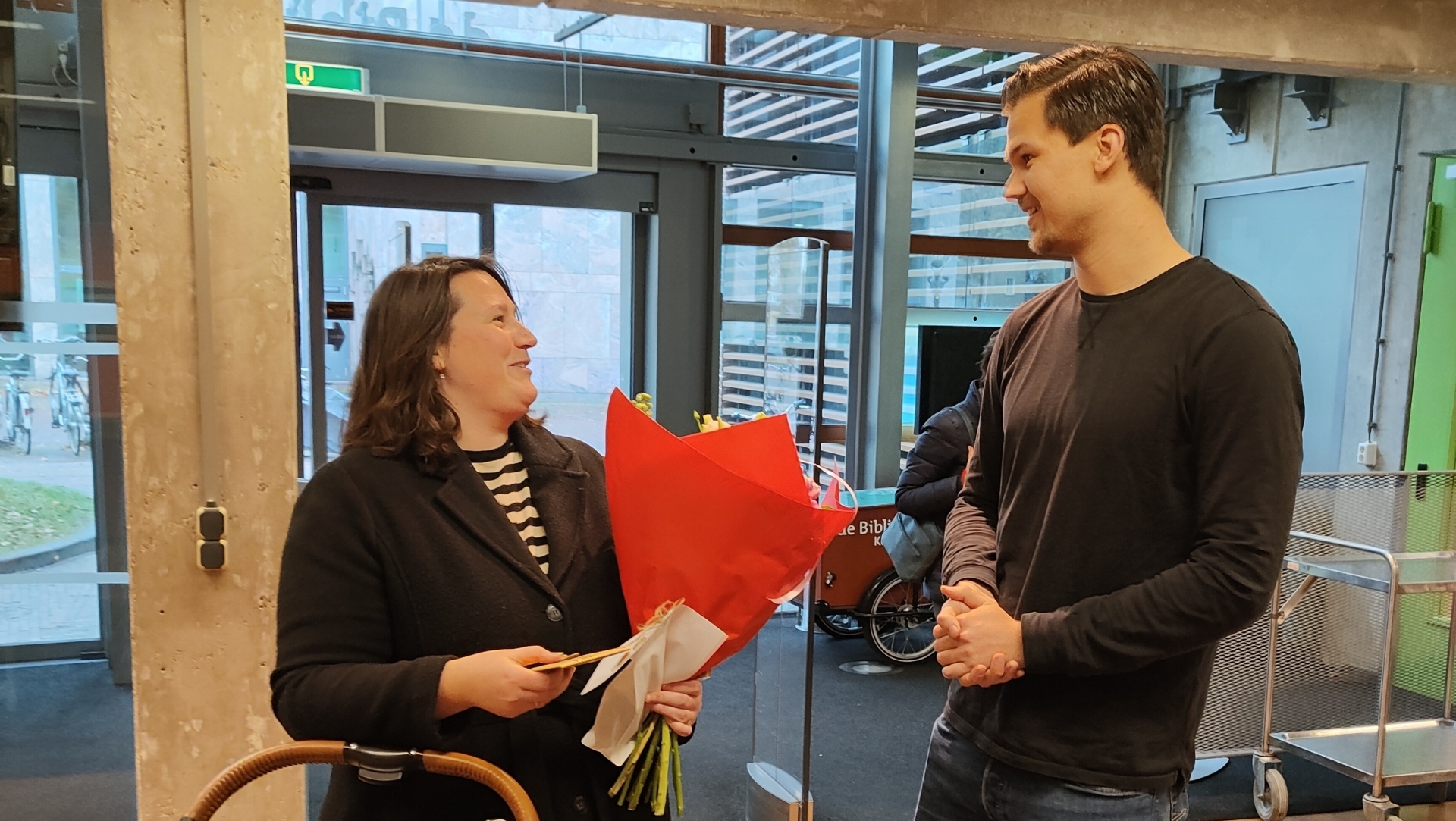 Een vrouw met een kinderwagen ontvangt een boeket bloemen van een man in een gebouw.