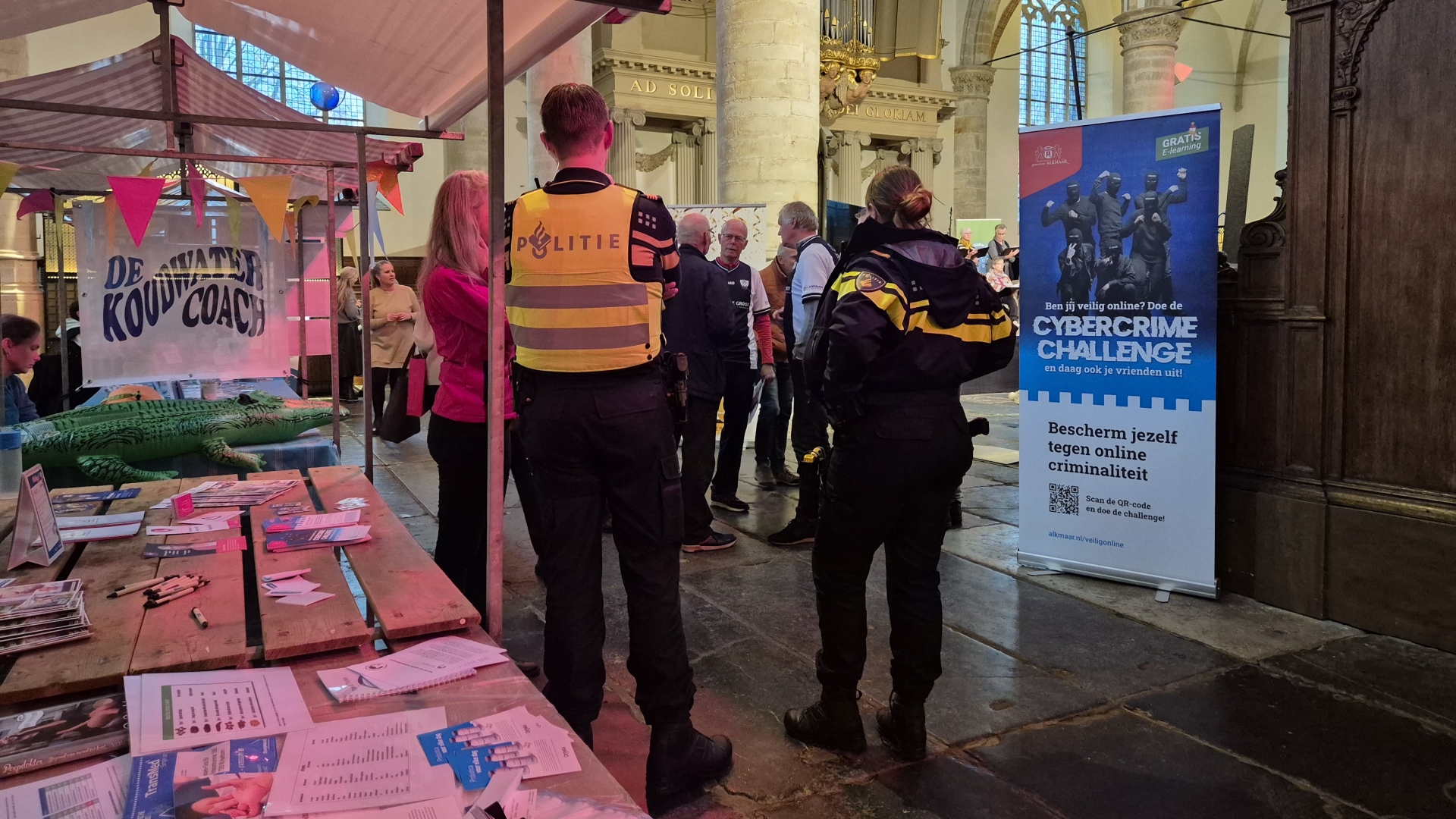 Een informatiestand met politieagenten en een poster voor een cybercrime-uitdaging in een hal met mensen en kleurige vlaggetjes.