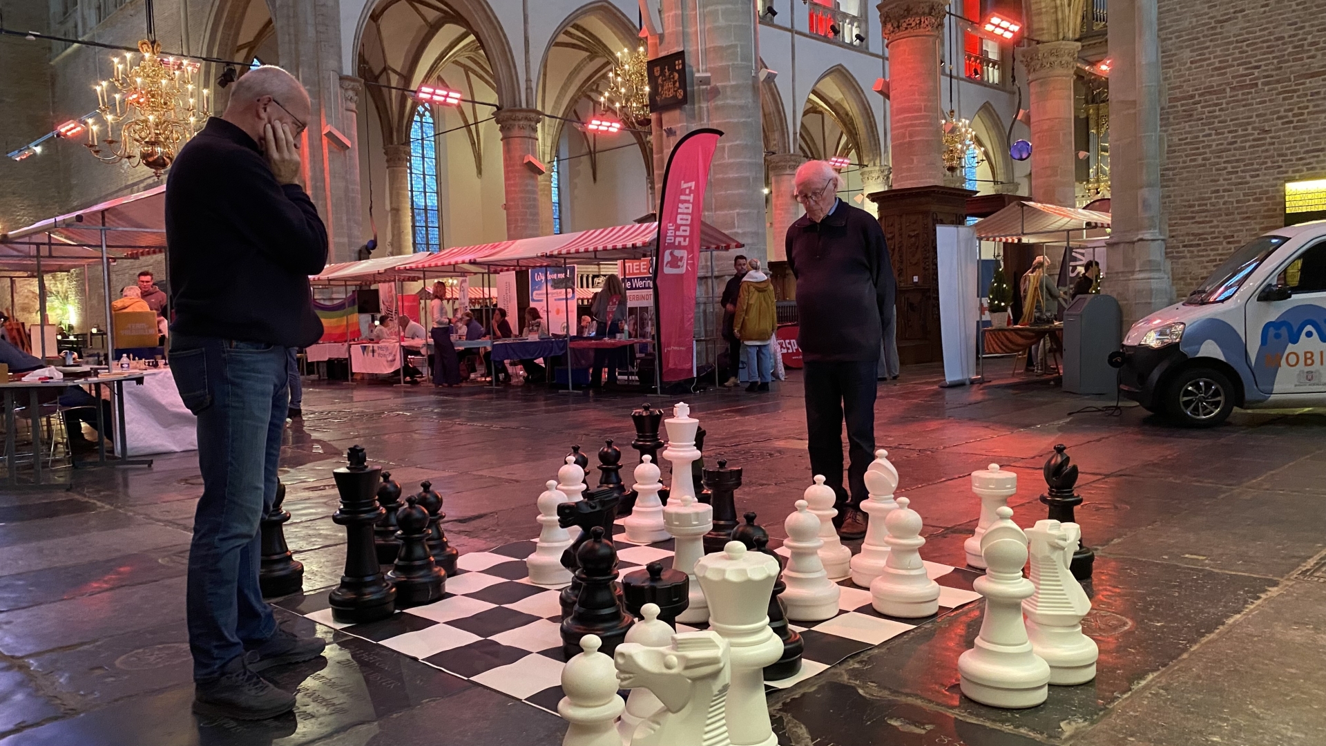 Twee mannen spelen schaak met grote schaakstukken in een kerk, met kraampjes en een bestelwagen op de achtergrond.