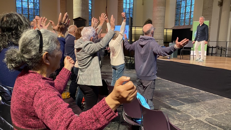 Mensen staan met opgeheven armen in een zaal bij een persoon op het podium.