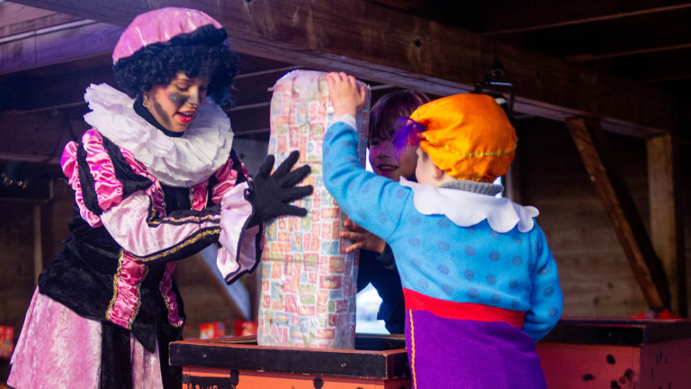 Twee kinderen in kleurrijke kostuums helpen een persoon in een Piet-outfit met een groot ingepakt cadeau.