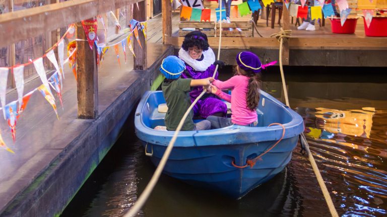 Kinderen in een boot met een persoon verkleed als Zwarte Piet in een kleurrijke omgeving met vlaggetjes.