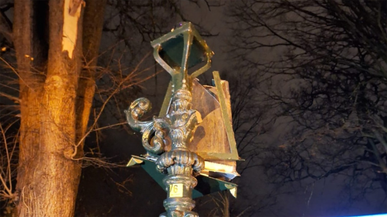 Beschadigde lantaarnpaal met gebroken onderdelen, omringd door kale bomen in de nacht.