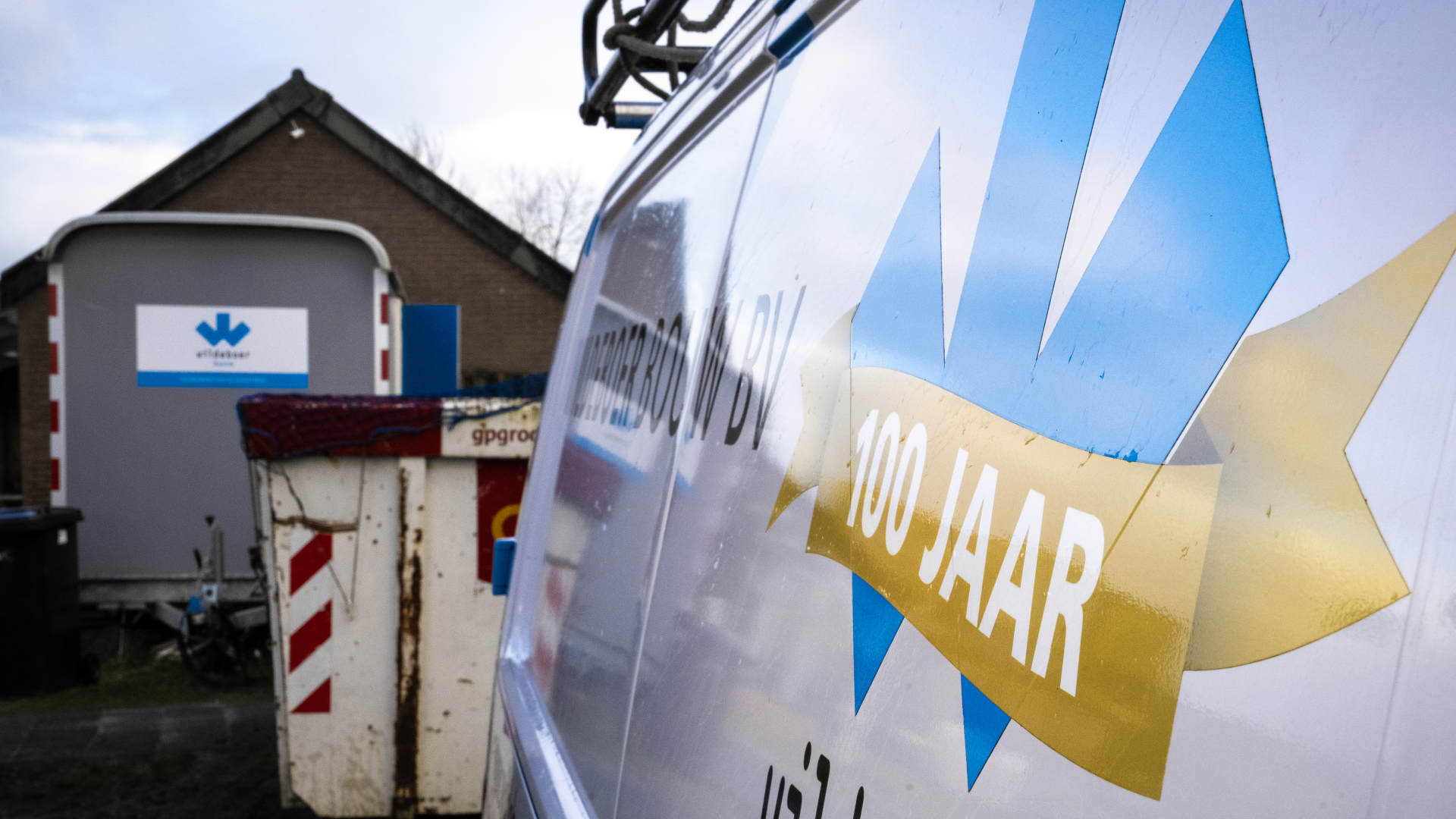 Een voertuig van een bouwbedrijf met het opschrift "100 jaar", naast een container en een gebouw met een bord erop.