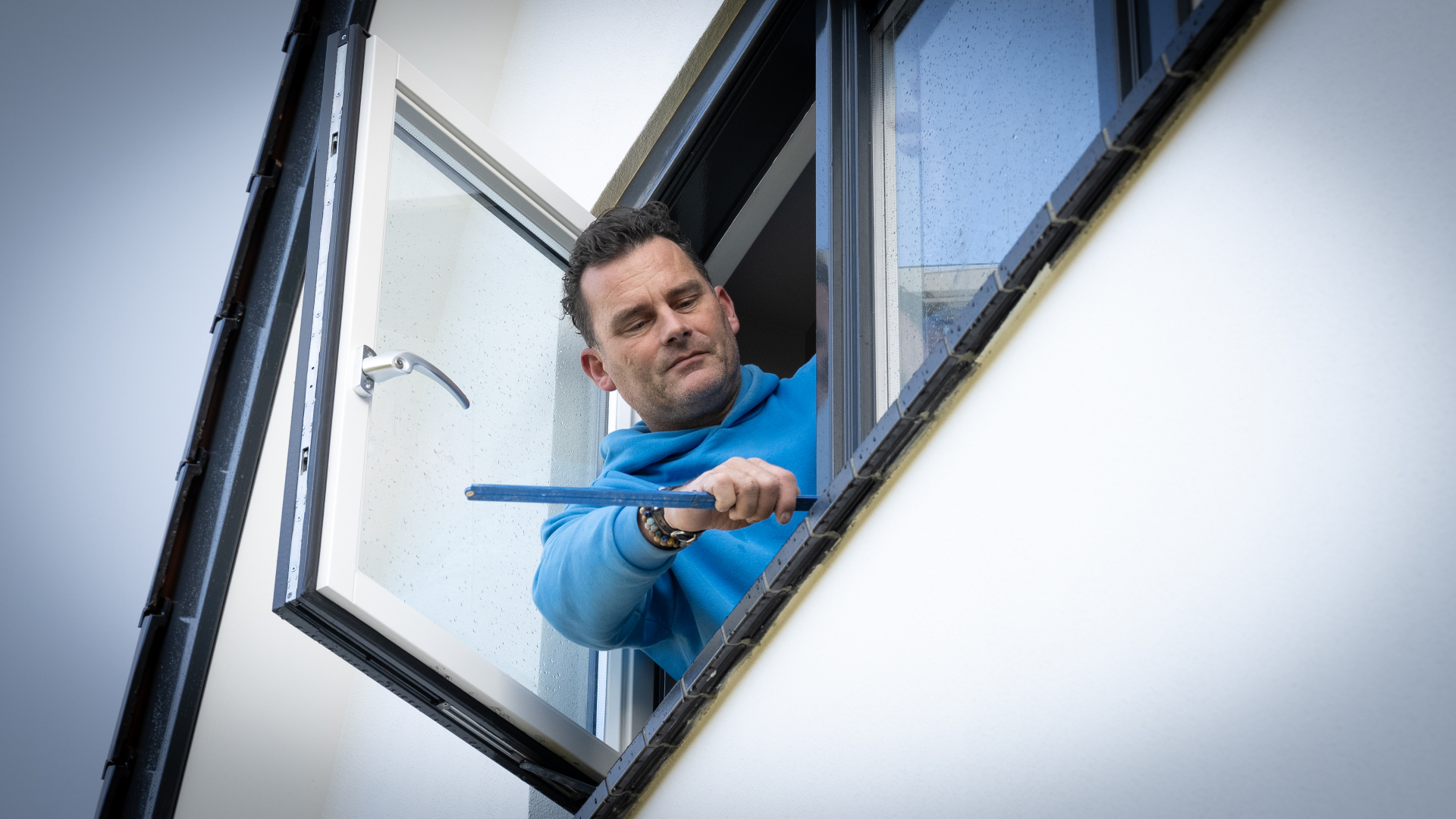 Man in een blauwe trui meet de buitenrand van een raam vanuit een openstaand raam.