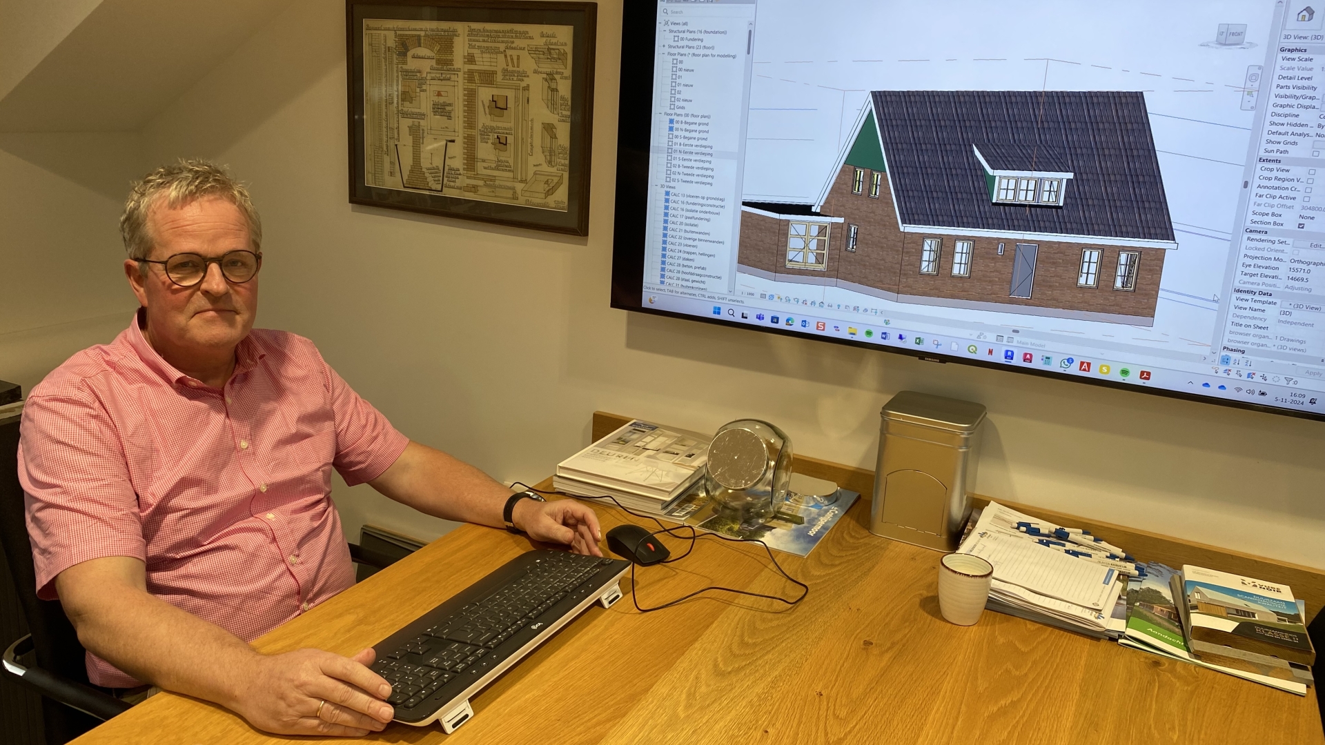 Man in een roze shirt zit aan een bureau met een computer, met een architecturaal ontwerp van een huis op een groot scherm.