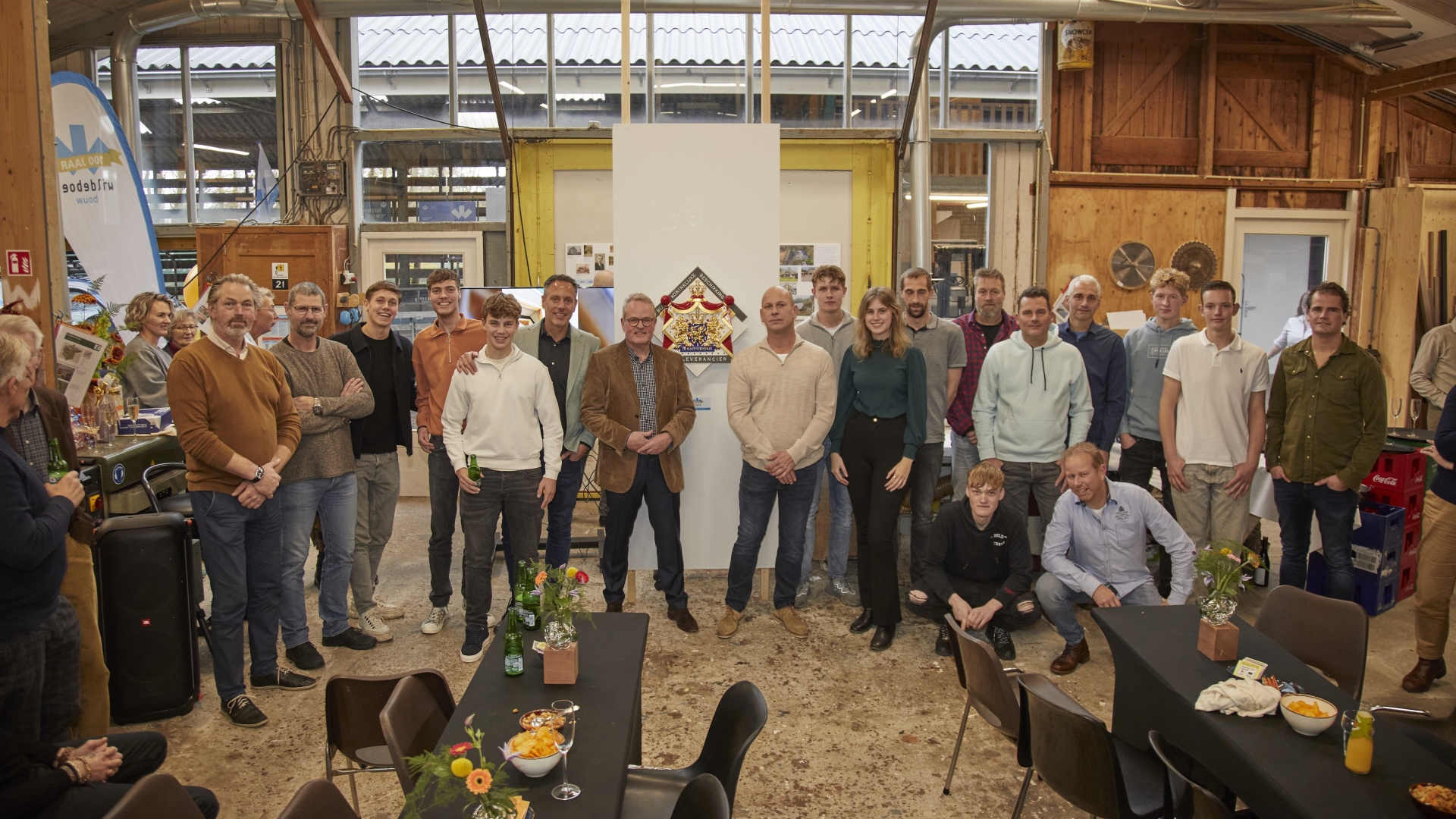 Een groep mensen poseert voor een foto in een werkplaats met tafels en bloemen.