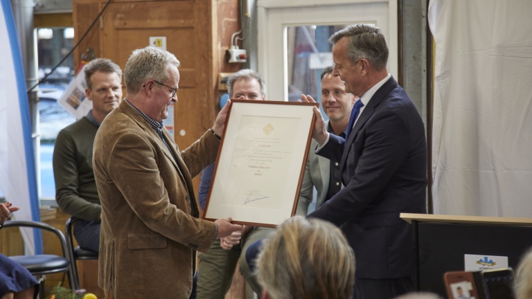 Twee mannen poseren met een ingelijste oorkonde, terwijl mensen op de achtergrond toekijken.