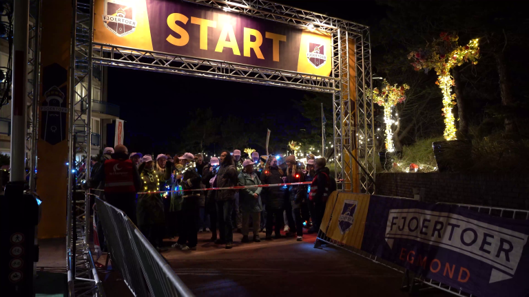 Mensen staan 's nachts onder een verlicht "START"-bord voor een evenement genaamd Fjoertoer in Egmond.