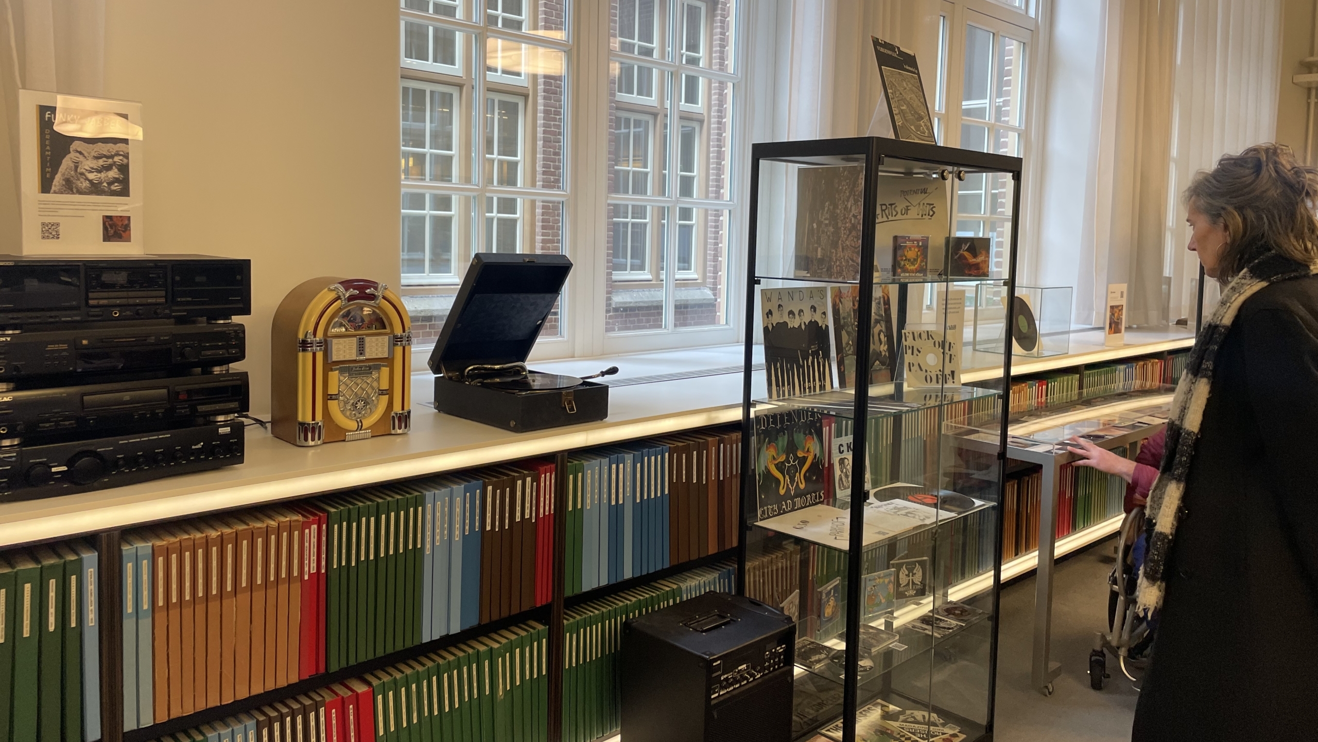 Vrouw bekijkt een vitrine met platen in een kamer met boekenplanken, een jukebox en een platenspeler.