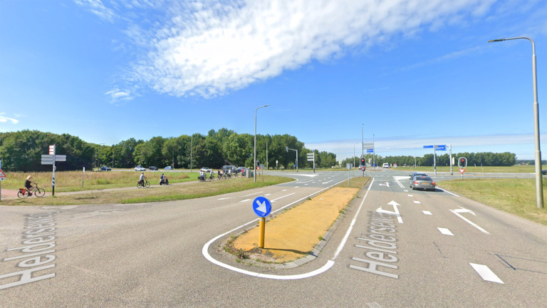 Wegkruising met fietspad, auto’s en fietsers, omgeven door grasvelden en bomen.