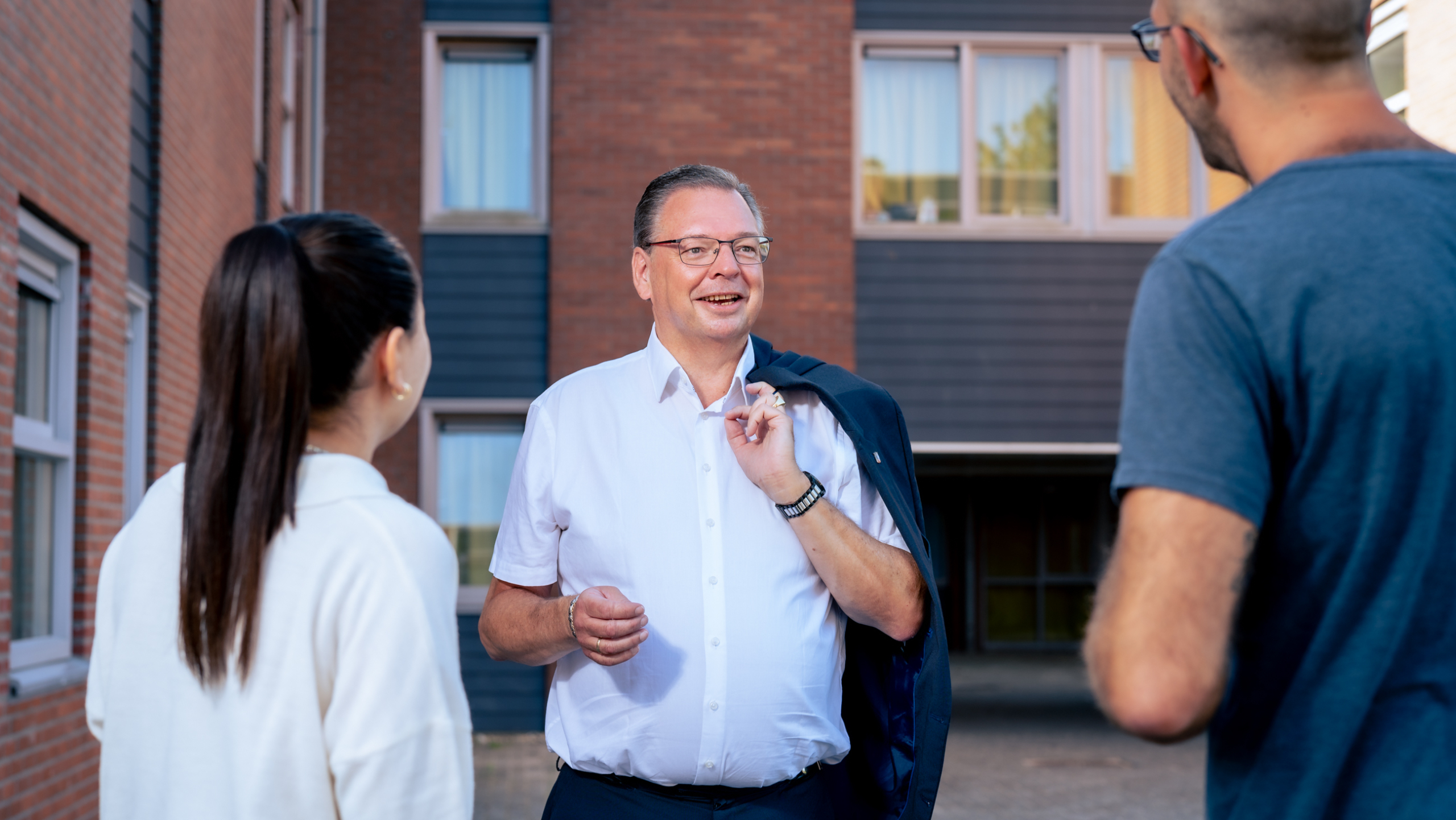 Drie mensen staan buiten en praten met elkaar voor een bakstenen gebouw.