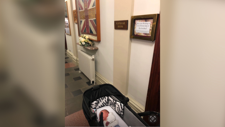 Een baby slaapt in een kinderwagen in een gang met een bord "Alkmaar Room", bloemen en een Britse vlag.