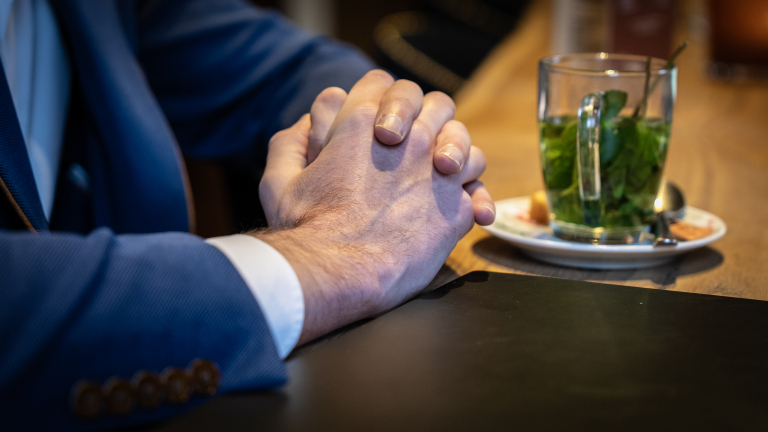 Man in blauw pak met gevouwen handen naast een glas muntthee op een houten tafel.