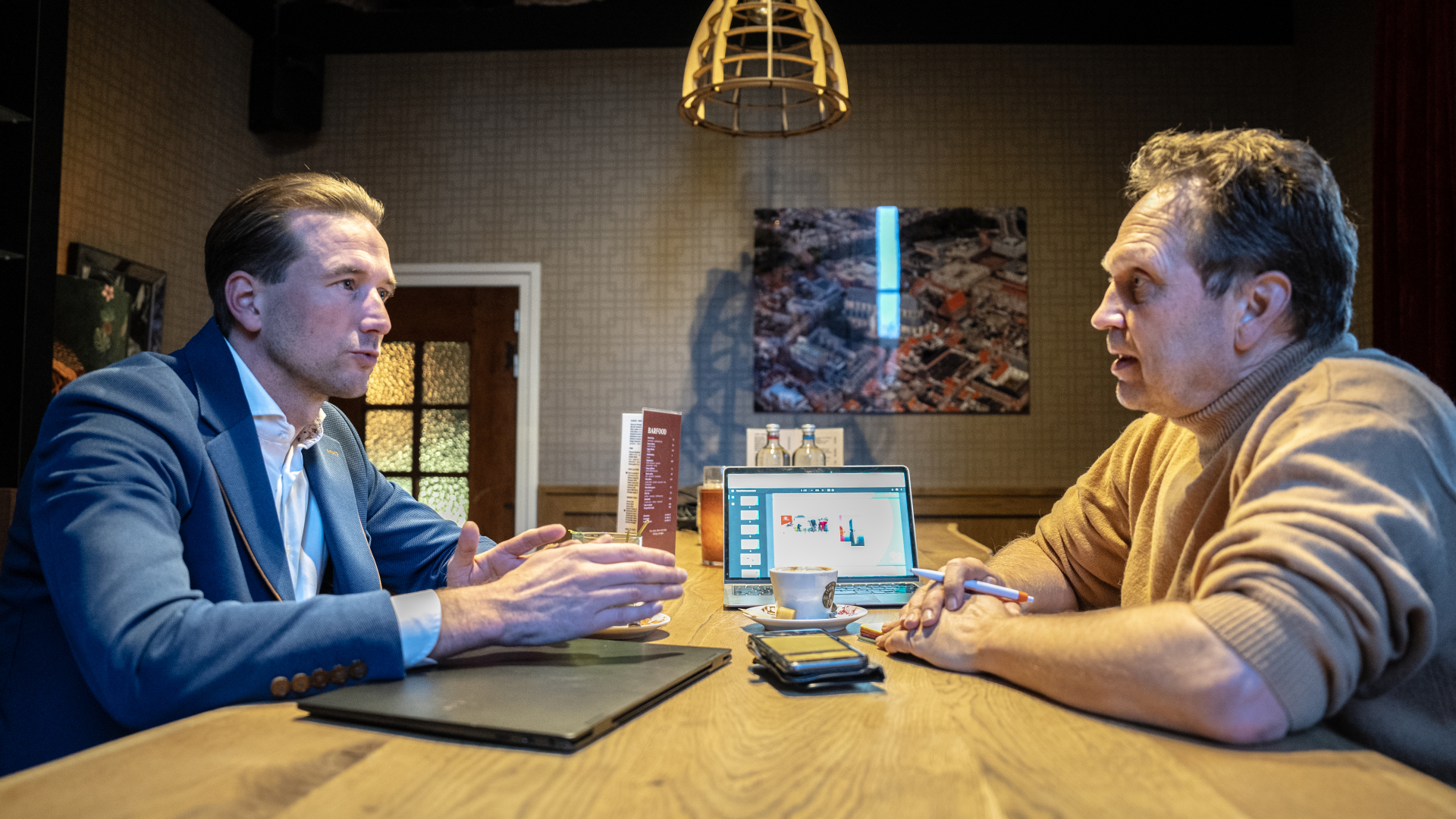 Twee mannen in gesprek aan een tafel met een laptop en koffiekopjes.