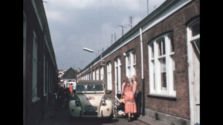 Straatbeeld met een klassieke auto geparkeerd naast een rij bakstenen huizen en mensen die converseren.