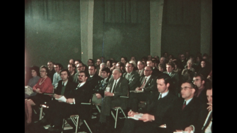 Een groep mensen zit in een zaal en luistert naar een presentatie.