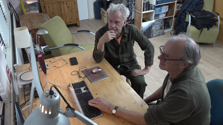 Twee mannen zitten aan een tafel met een computer in een kantooromgeving.