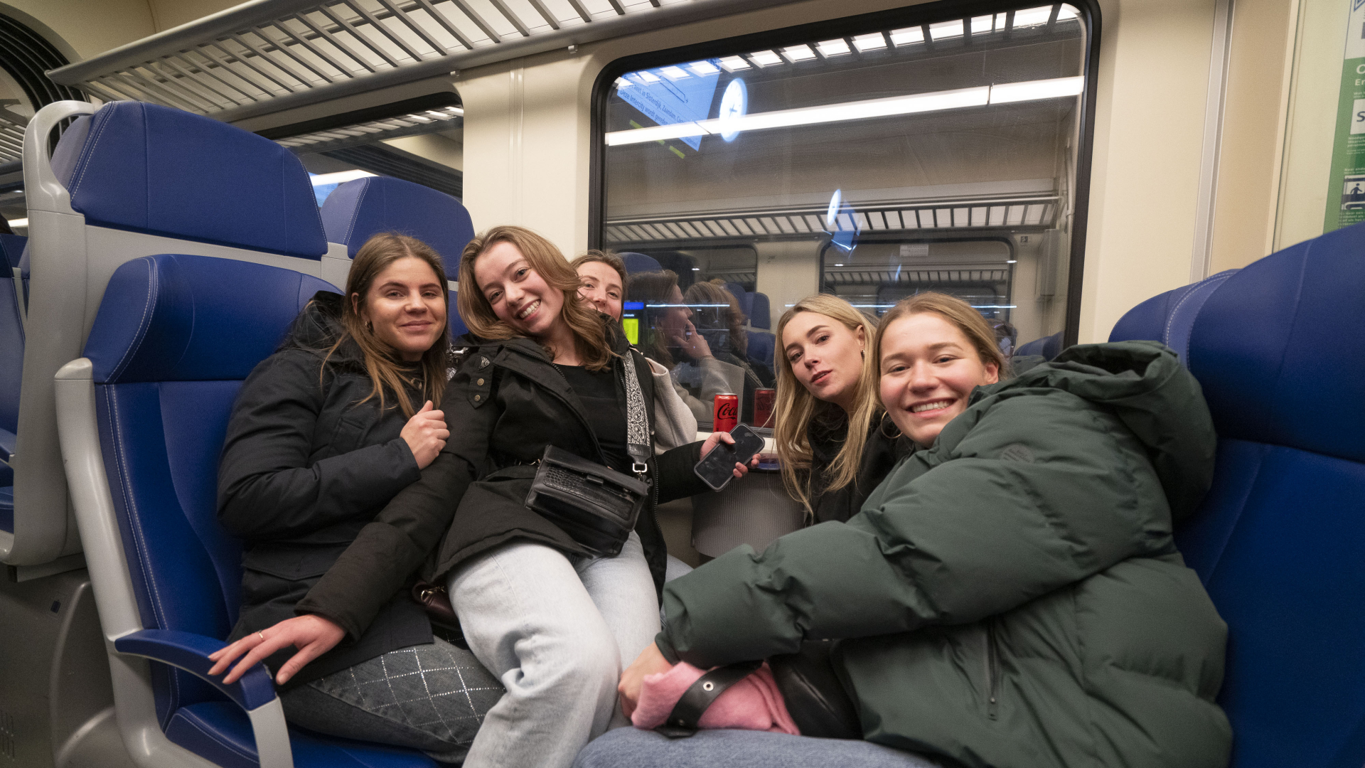 Groep jonge vrouwen zit samen in een trein, glimlachend naar de camera.