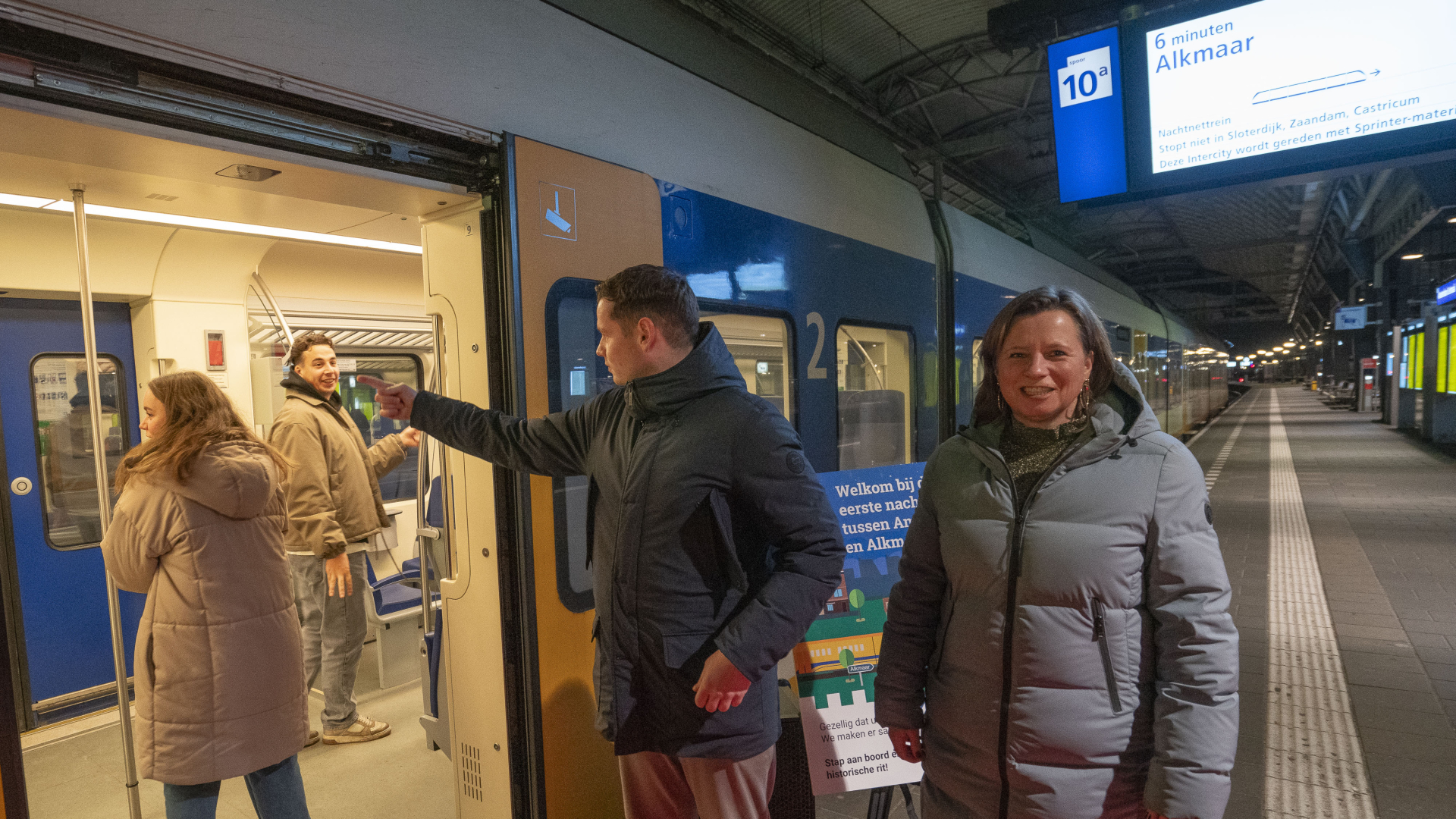 Mensen stappen in een trein op een station in de nacht; een bord toont de bestemming Alkmaar.