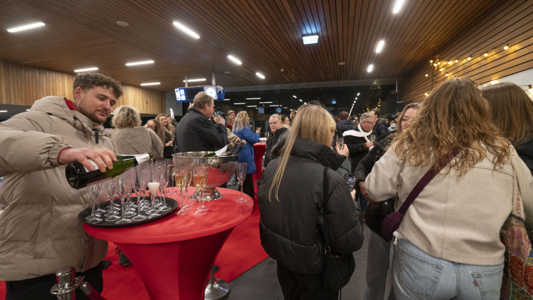 Mensen verzamelen zich in een ruimte met een houten plafond, terwijl een man champagne inschenkt in glazen op een rode tafel.