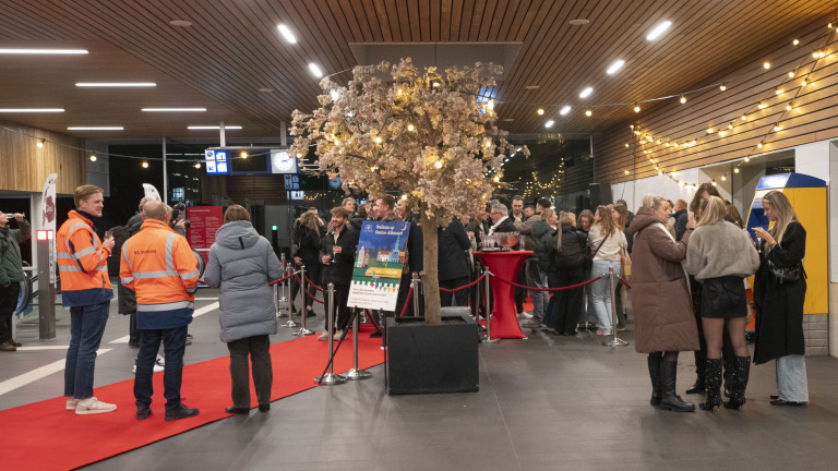 Mensen verzamelen zich bij een versierd station met rode loper en feestelijke verlichting.