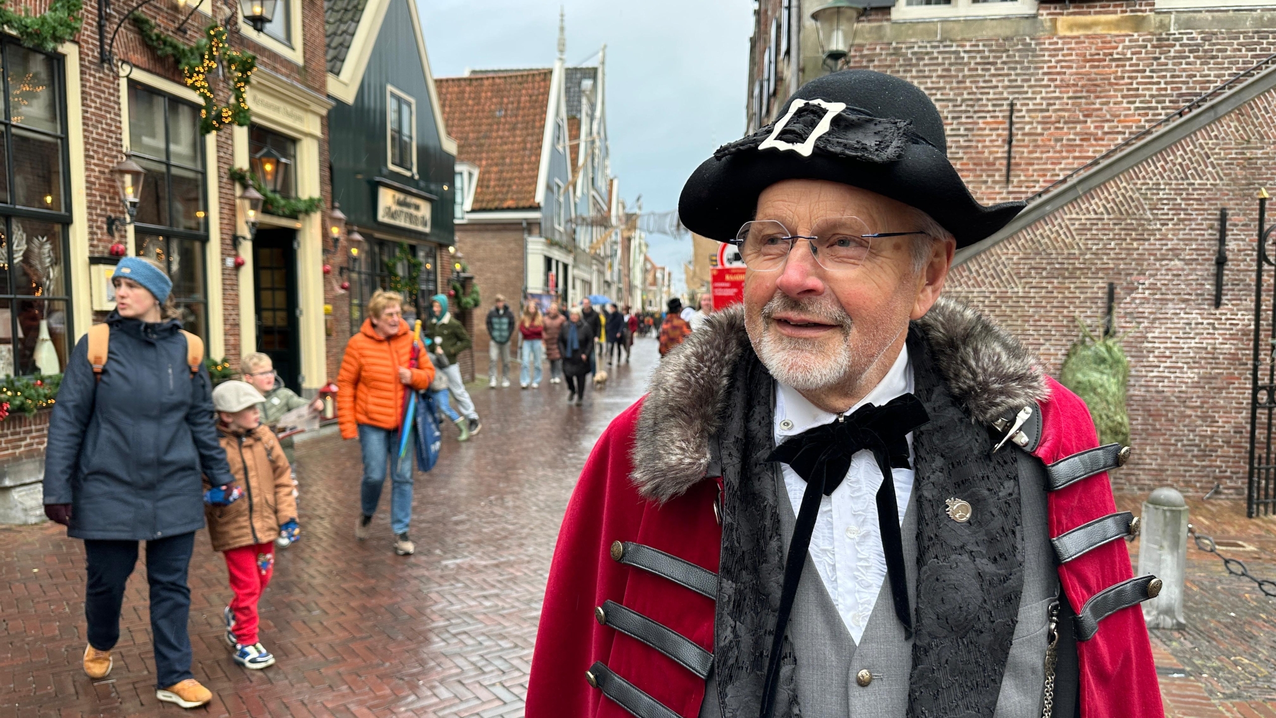 Een oudere man in historische kleding met een zwarte hoed en rode mantel loopt door een drukke winkelstraat.