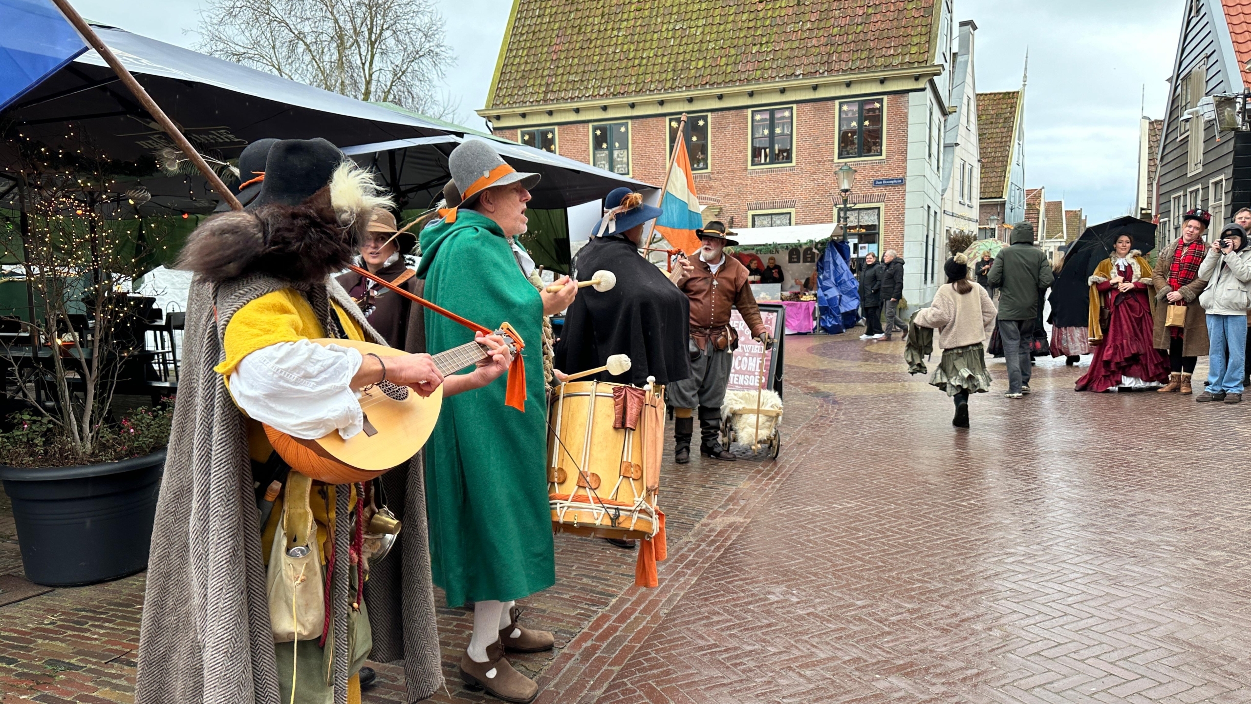 Mensen in historische kleding spelen muziek op een straatmarkt met kasseien, omgeven door oude gebouwen.