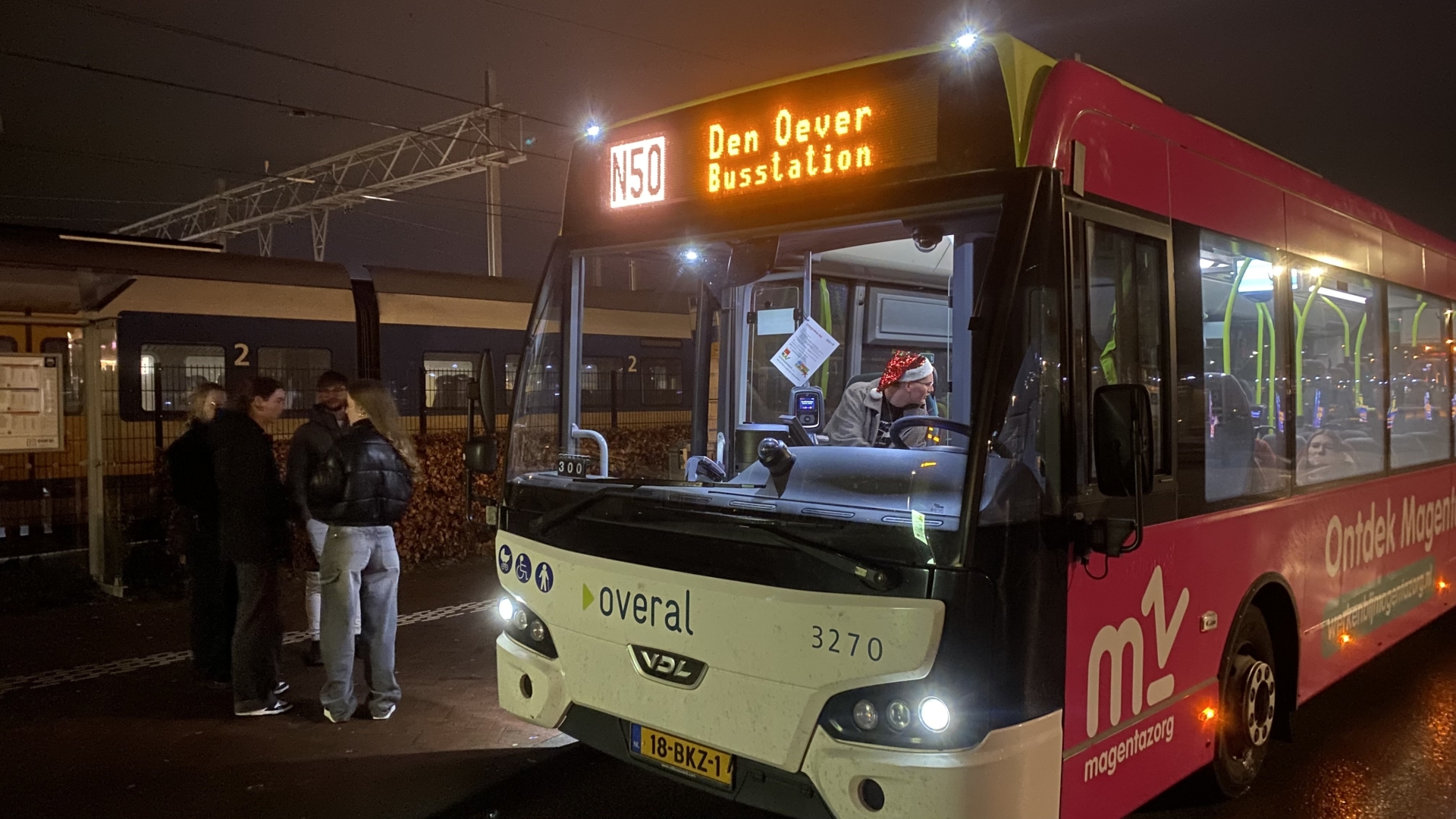 Een bus met de tekst "N50 Den Oever Busstation" op het display staat bij een halte in het donker. Naast de bus staan enkele mensen en een persoon met een kerstmuts achter het stuur.