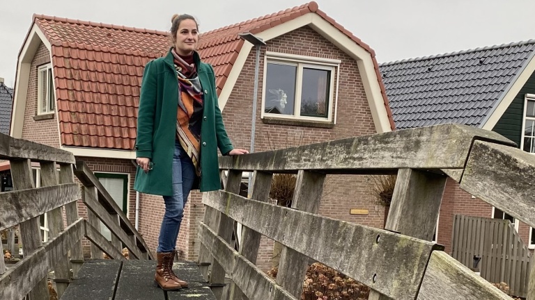 Vrouw in groene jas op houten brug met huizen op de achtergrond.