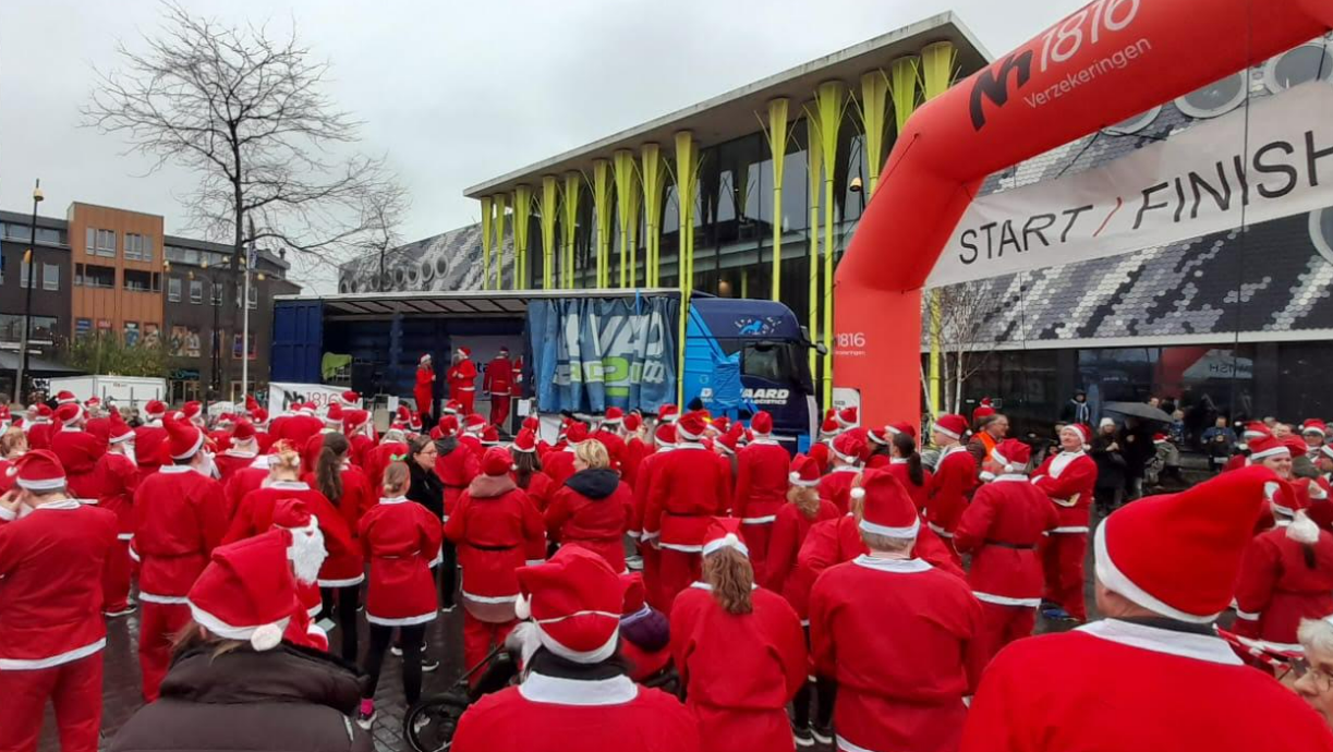 Mensen in kerstmannenpakken verzamelen zich bij een start/finishlijn voor een evenement. Op de achtergrond staat een vrachtwagen.